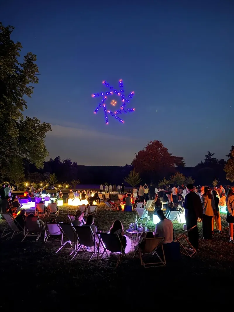 Figure violette et orange dessinée par les drones au crépuscule, devant les invités